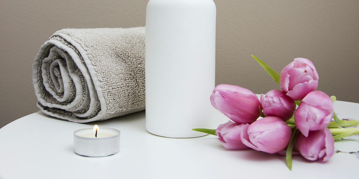 Une serviette éponge roulée, un flacon d’huile sans étiquette et une bougie allumée posés sur une table blanche